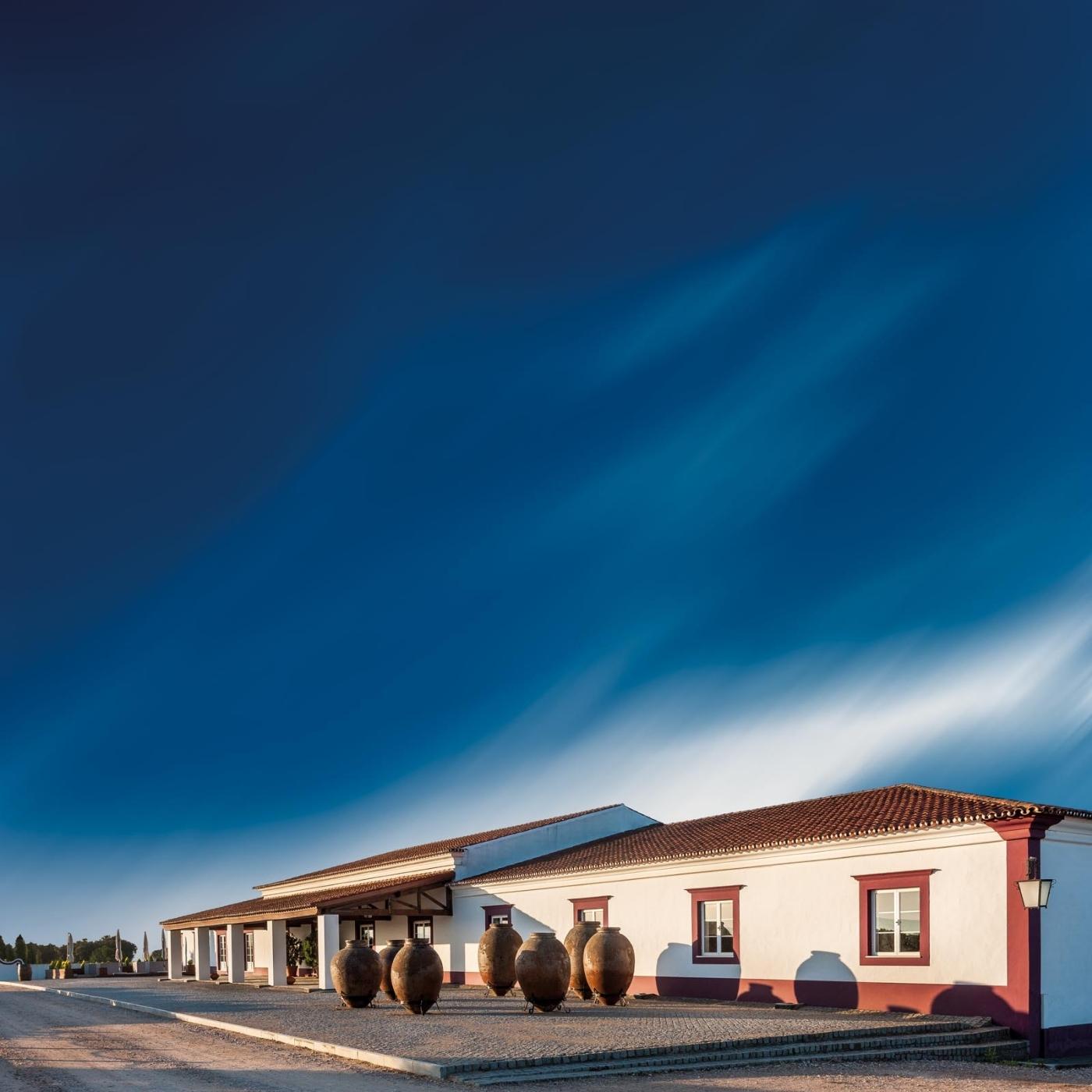 Erlesene Weine vom Weingut HERDADE DAS SERVAS aus Portugal/Alentejo. Bild vom Weingut, weiß-rotes Gebäude mit kleinen Bäumen an einer Straße, oberhalb ein azurblauer Himmel.