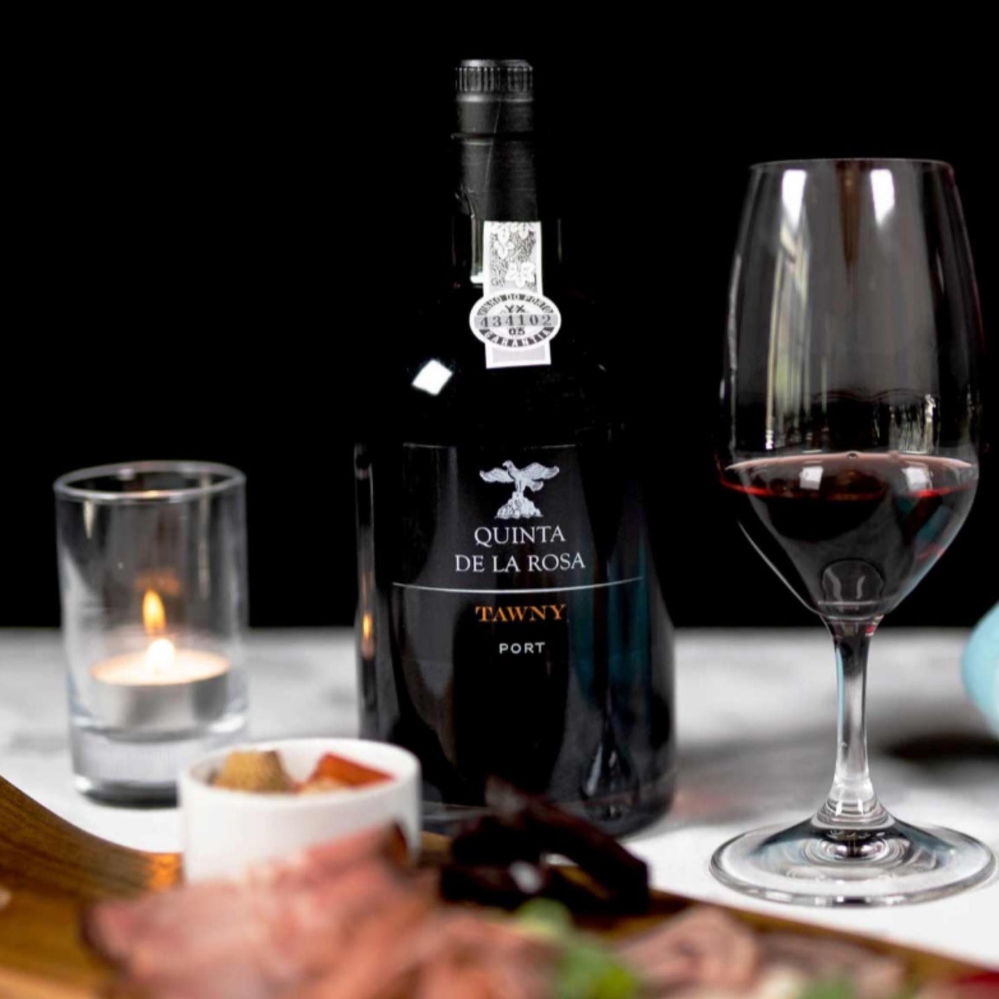 
                  
                    TAWNY PORT, Portwein vom Weingut Quinta de la Rosa. Bild mit Portweinflasche und Portweinglas, daneben eine brennende Kerze und portugiesische Speisen.
                  
                