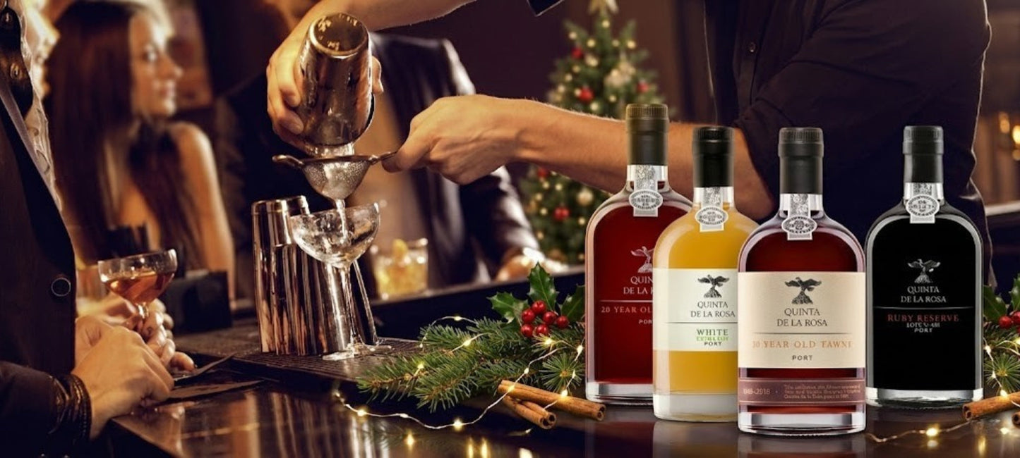 Barkeeper gießt einen Drink in einer weihnachtlich dekorierten Bar ein; im Vordergrund stehen vier Flaschen Quinta de la Rosa Portwein (20 Year Old, White, Tawny und Ruby Reserve) dekoriert mit Tannenzweigen, Zimtstangen und Lichterketten.