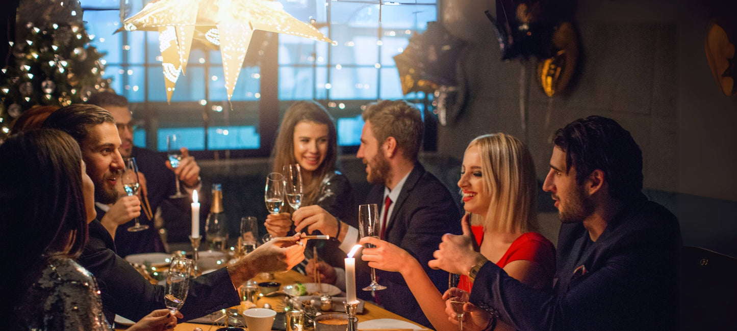 Junge Menschen sitzen am Tisch, essen und trinken und feiern gemeinsam Weihnachten. Im Hintergrund ist ein Christbaum und ein Weihnachtsstern zu sehen.