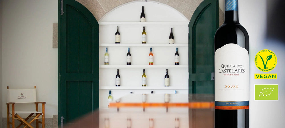 Eine Flasche Rotwein der Marke Quinta dos Castelares mit veganen und Bio-Siegeln steht vor einem Regal mit weiteren Weinflaschen in einem Eingangsbereich.