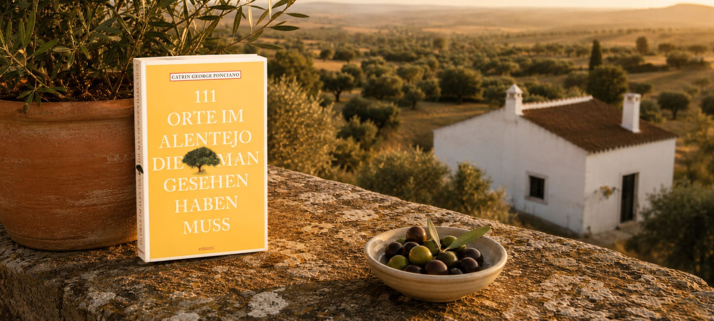 Gelbes Buch mit dem Titel „111 Orte im Alentejo...“ lehnt an einem Tontopf auf einer Steinmauer neben einer Schale Oliven, im Hintergrund ein weißes Landhaus in einer mediterranen Hügellandschaft bei Sonnenuntergang.