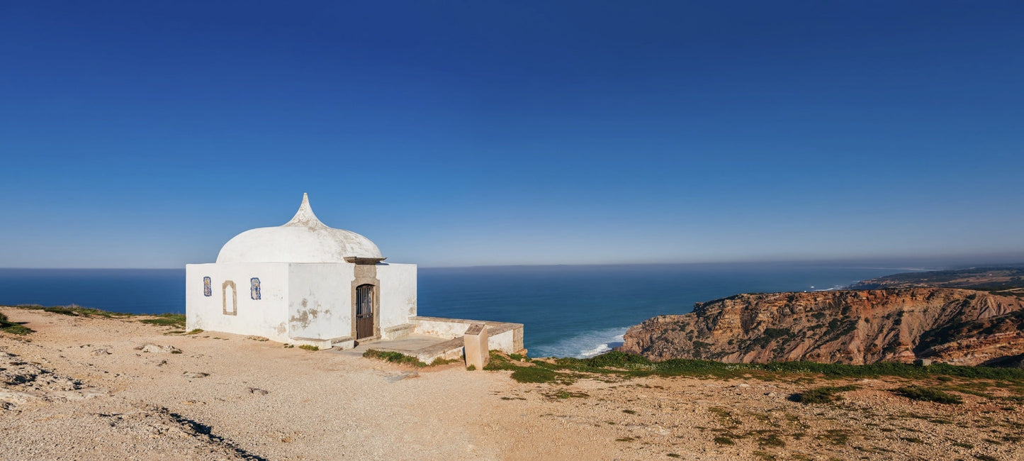 Kleine weiße Kapelle mit markanter, geschwungener Kuppel direkt am Rand einer Steilküste vor dem blauen Atlantik am Cabo Espichel, Portugal.