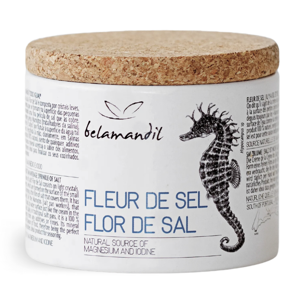 Ein runder Behälter mit Korkdeckel, etikettiert als “Fleur de Sel Flor de Sal”. Das Etikett beschreibt das Produkt als natürliches Meersalz, reich an Mineralien wie Magnesium und Jod. Eine Illustration eines Seepferdchens symbolisiert die Herkunft des Salzes aus dem Meer