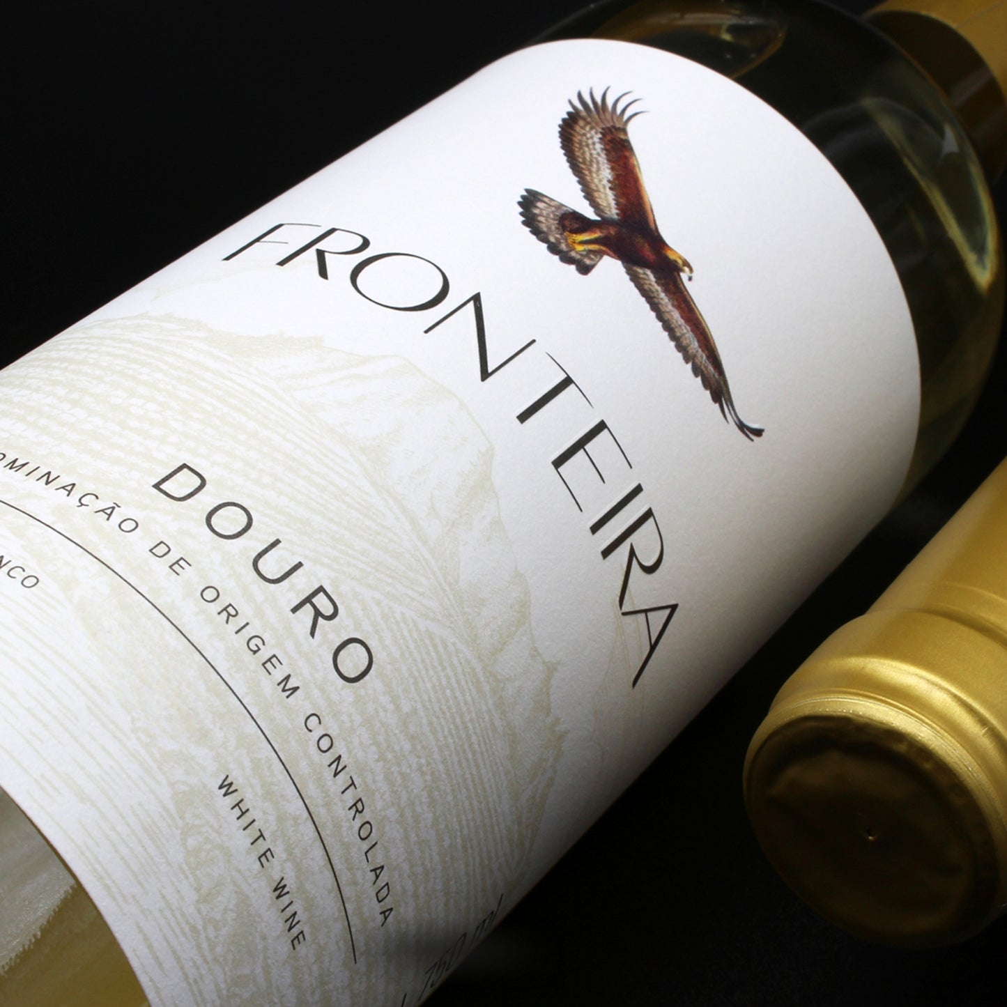 Flasche Fronteira Colheita Weißwein aus dem Douro-Gebiet, Portugal. Das Etikett ist weiß und zeigt einen Adler im Flug über einer Berglandschaft, darüber der Schriftzug „FRONTEIRA“ und darunter „DOURO“. Der Wein ist hellgelb und die Flasche hat einen goldenen Schraubverschluss.