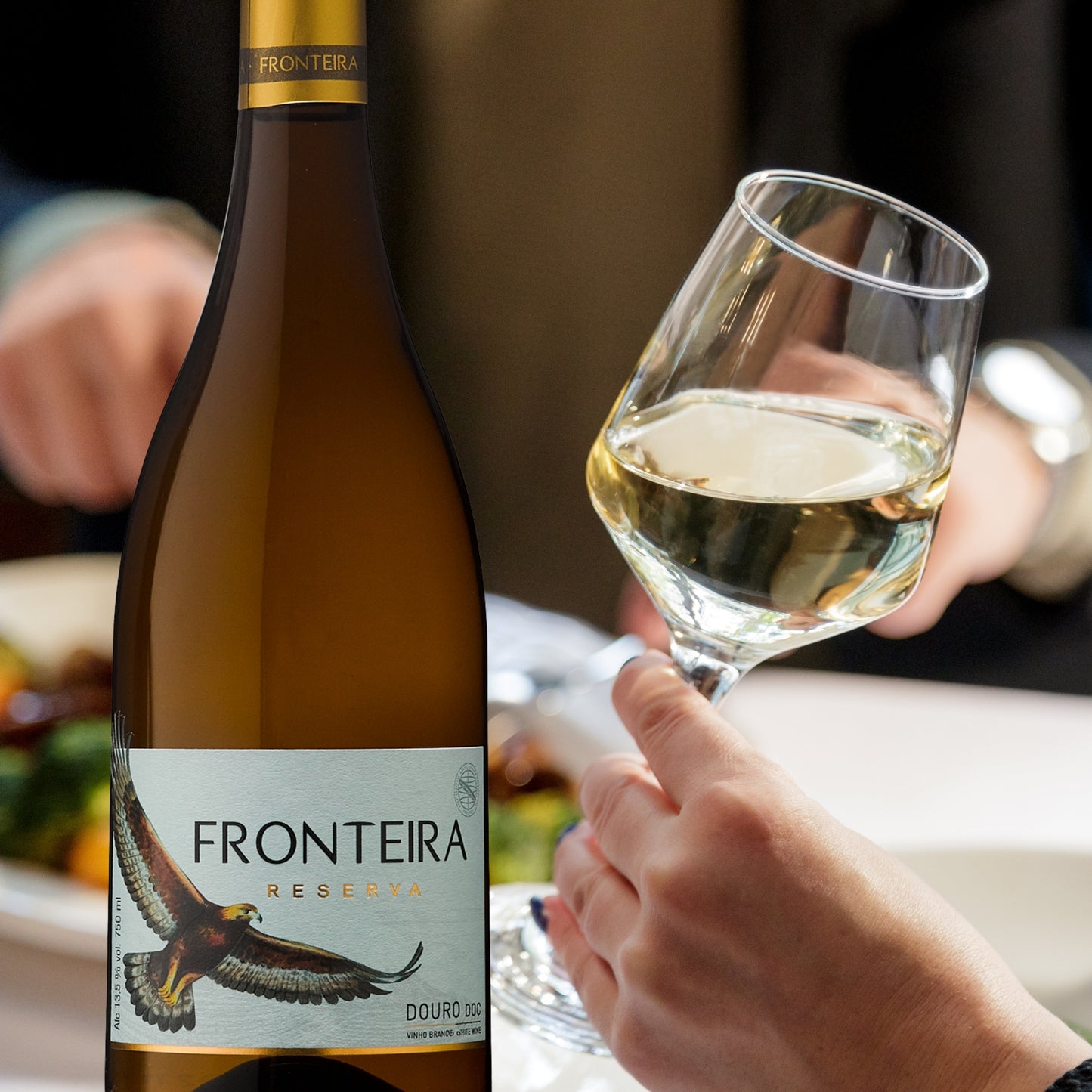 Flasche Fronteira Reserva Weißwein aus dem Douro-Gebiet, Portugal. Der Wein ist dunkelgelb/bernsteinfarben und die Flasche ist bauchiger als das Standardmodell. Das Etikett ist weiß und zeigt einen goldbraunen Adler, der auf die Seitenkante blickt, darüber der Schriftzug „FRONTEIRA RESERVA“ und darunter „DOURO DOC“. Die Flasche hat einen goldenen Schraubverschluss.