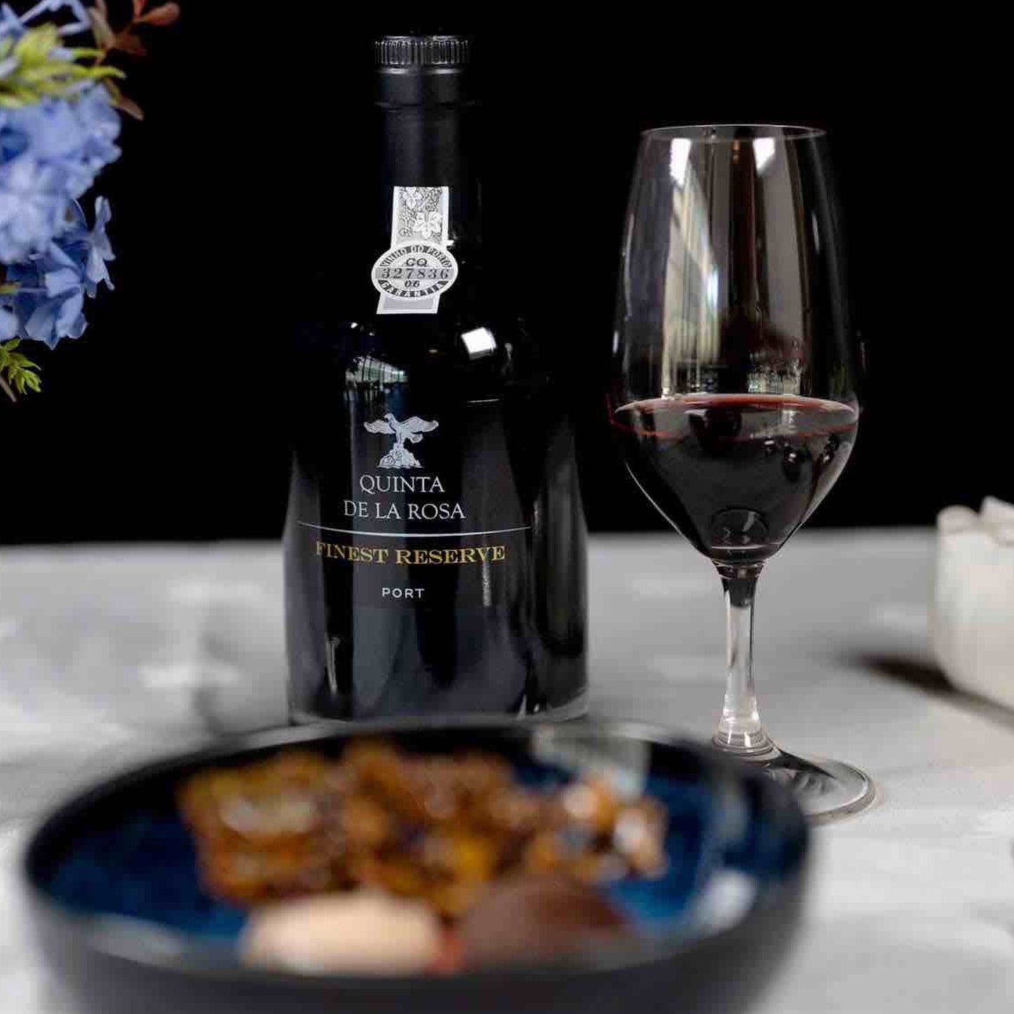 
                  
                    Eine Flasche Quinta de la Rosa Finest Reserve Port und ein gefülltes Weinglas stehen neben einer Schale mit Vorspeisen auf einem elegant gedeckten Tisch.
                  
                