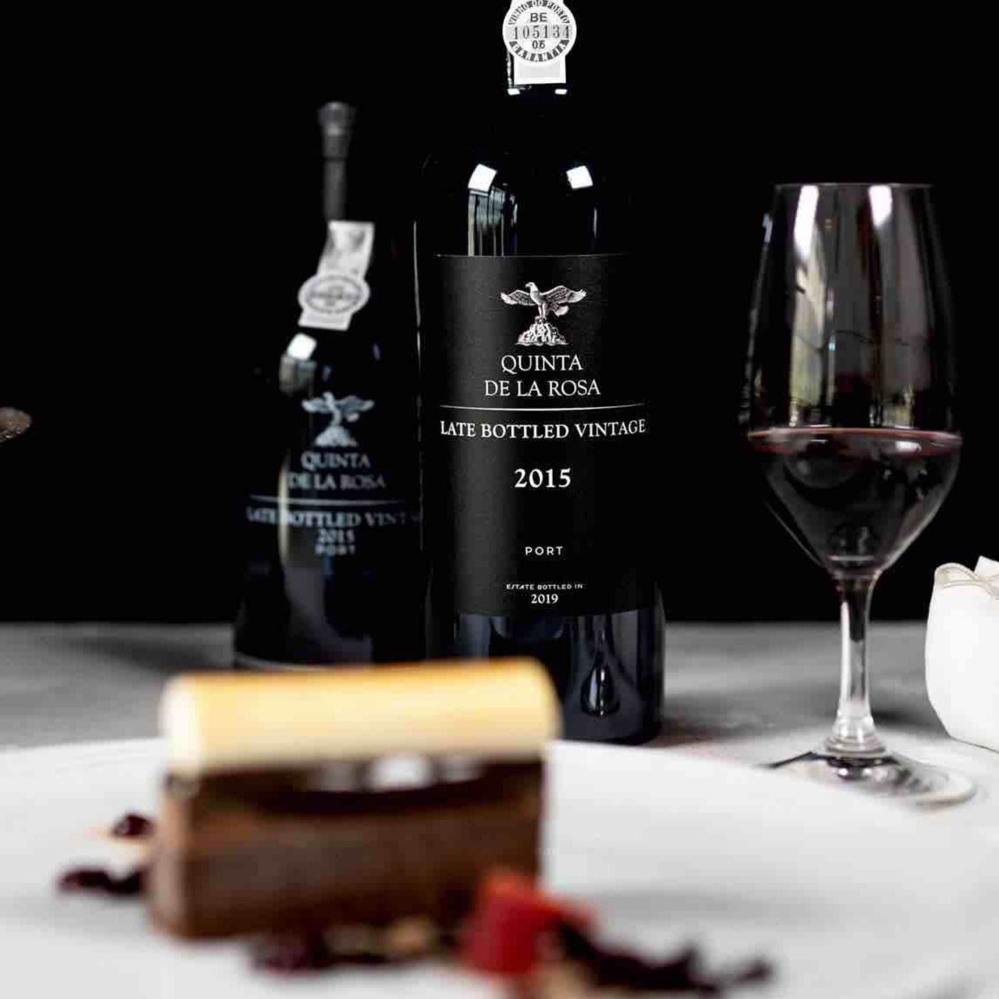 Arrangement von zwei Flaschen Quinta de la Rosa Late Bottled Vintage Port 2015, einem gefüllten Weinglas und einem kunstvoll angerichteten Dessert, perfekt für einen genussvollen Abend.