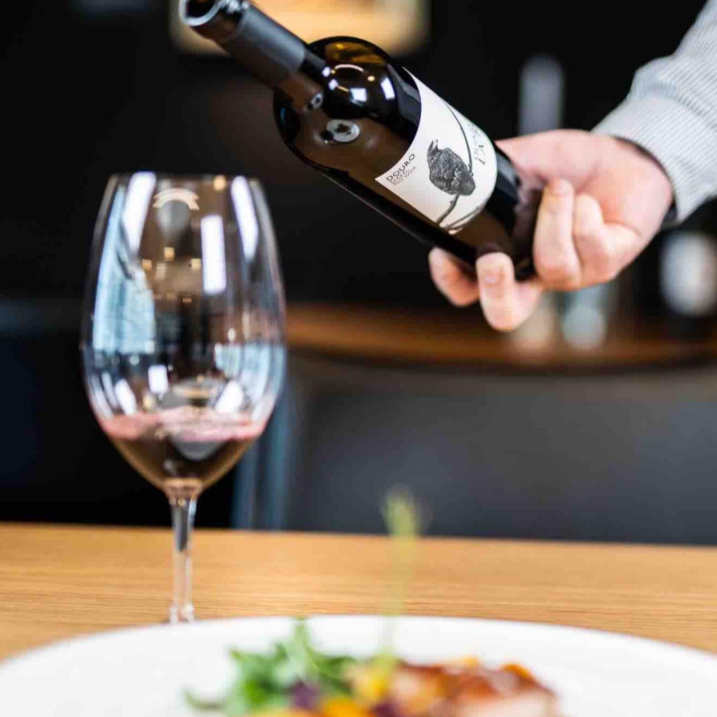 
                  
                    QUINTA DE LA ROSA ROTWEIN 2022 vom Weingut Quinta de la Rosa aus dem Douro/Portugal. Auf dem Bild ist ein Weinglas zu sehen, jemand schüttet Wein ein, davor steht ein Teller mit Essen.
                  
                