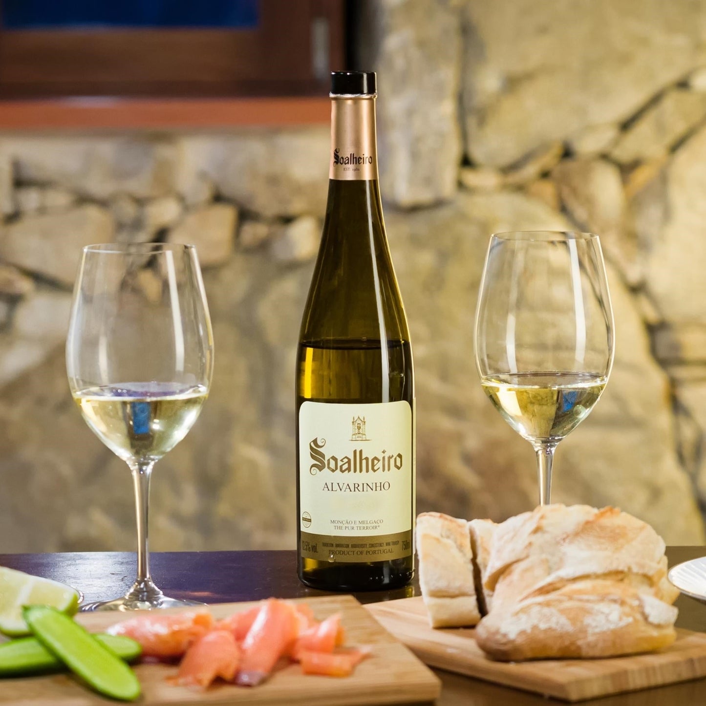 
                  
                    Alvarinho Classic, ein Weißwein vom Weingut Quinta da Soalheiro aus der Region Vinho Verde, Mino, Lima Tal, in Portugal. Bild mit Weinflasche und zwei Weißweingläsern auf einem Tisch stehend, daneben Holzbrettchen mit Weißbrot und Gemüse und Fisch.
                  
                