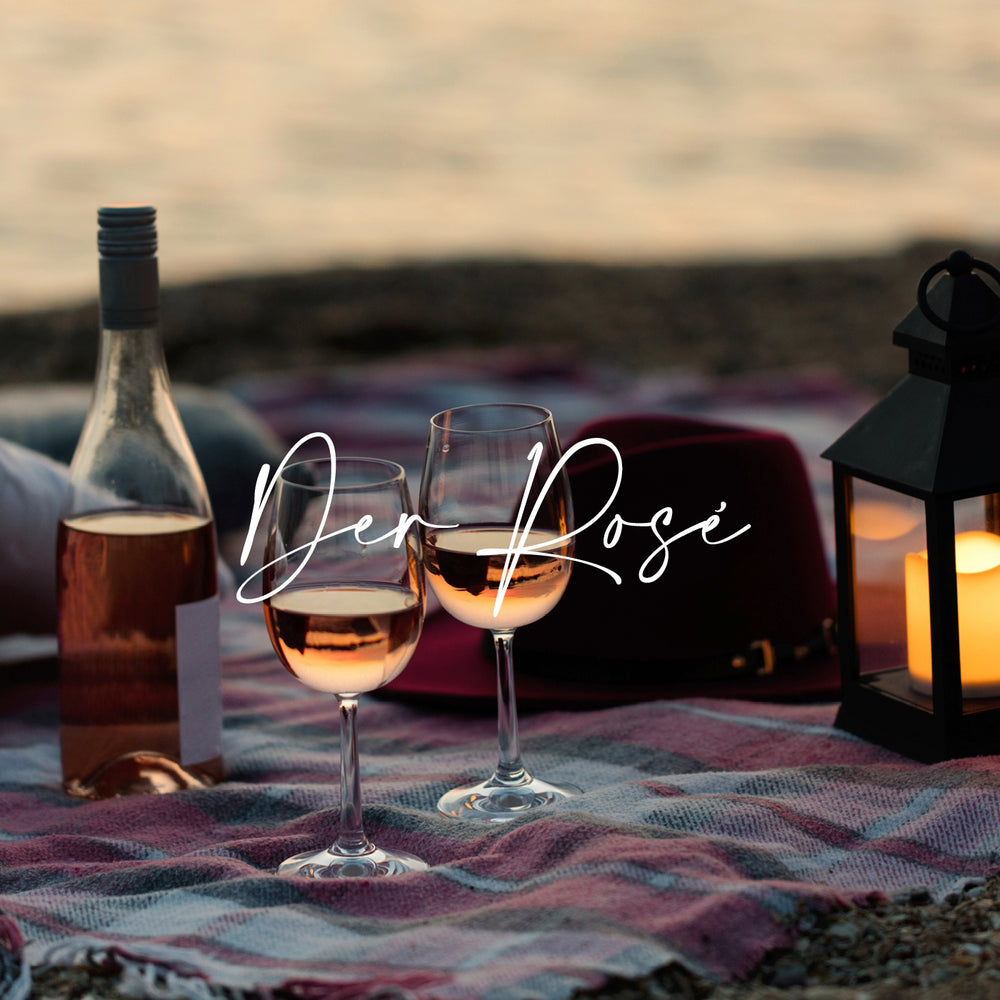 Roséweine von der Vinho Bar in Wuppertal. Bild am Strand: zwei Gläser mit Rosé gefüllt, daneben eine Rosé-Weinflasche und eine Laterne mit einer brennenden Kerze.