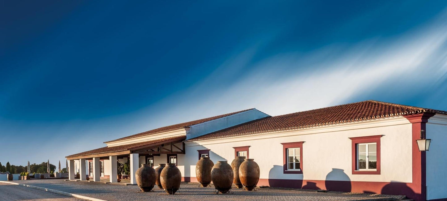 Erlesene Weine vom Weingut HERDADE DAS SERVAS aus Portugal/Alentejo. Bild vom Weingut, weiß-rotes Gebäude mit kleinen Bäumen an einer Straße, oberhalb ein azurblauer Himmel.