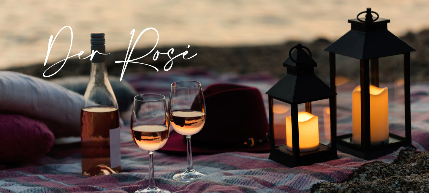Roséweine von der Vinho Bar in Wuppertal. Bild am Strand: zwei Gläser mit Rosé gefüllt, daneben eine Rosé-Weinflasche und eine Laterne mit einer brennenden Kerze.