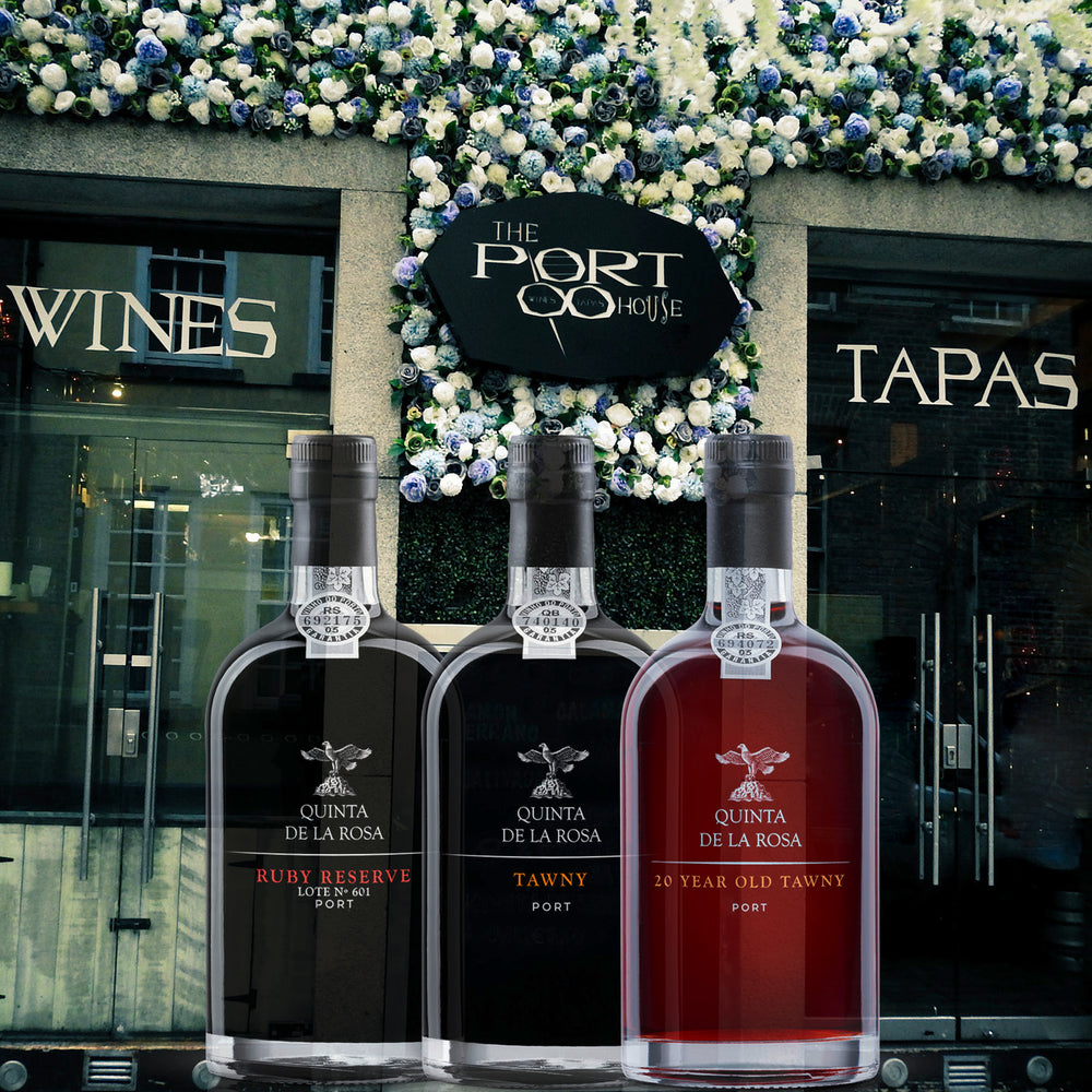 Das Foto zeigt drei Flaschen mit verschiedenen Portweinen von Quinta de la Rosa. Die Sorten umfassen Ruby, Tawny 10 Years und 20 Years. Im Hintergrund erstreckt sich eine malerische Weinregion in Portugal.