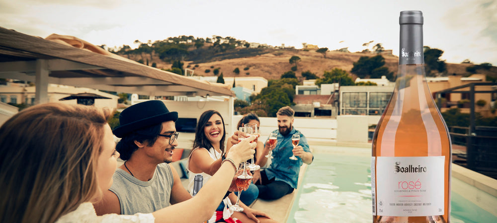 Das Foto zeigt eine Gruppe junger Menschen, die am Pool sitzen und Soalgeiro Alvarinho Rosé in eleganten Weingläsern genießen. Im Hintergrund erstreckt sich eine malerische mediterrane Landschaft mit charmanten Häusern.