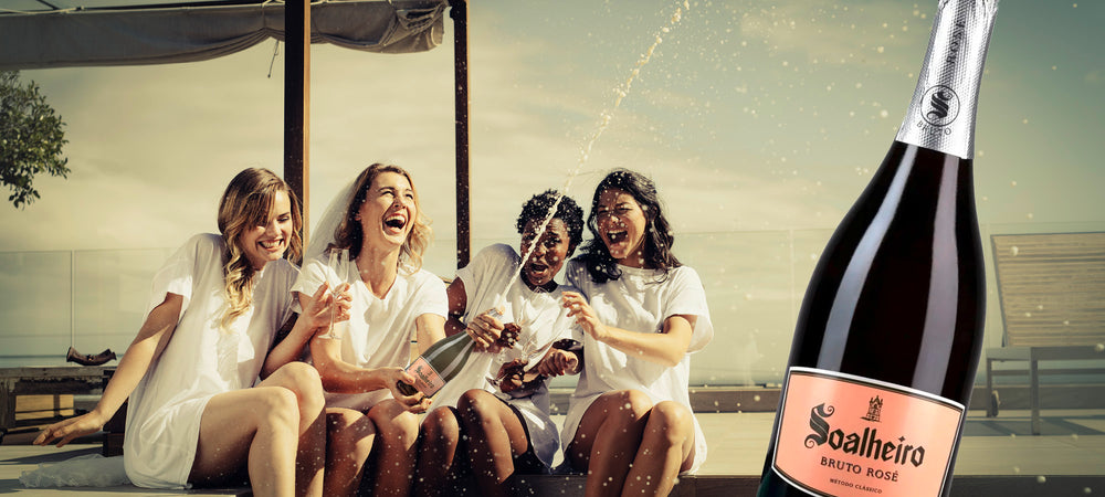 Ein Foto zeigt junge Frauen, die am Pool sitzen und eine Flasche Soalheiro Alvarinho Schaumwein Rosé geöffnet haben. Der Schaumwein spritzt aus der Flasche und sorgt für einen fröhlichen Moment. Es scheint, dass die Frauen einen Junggesellinnenabschied feiern und viel Spaß haben, während sie lachen.