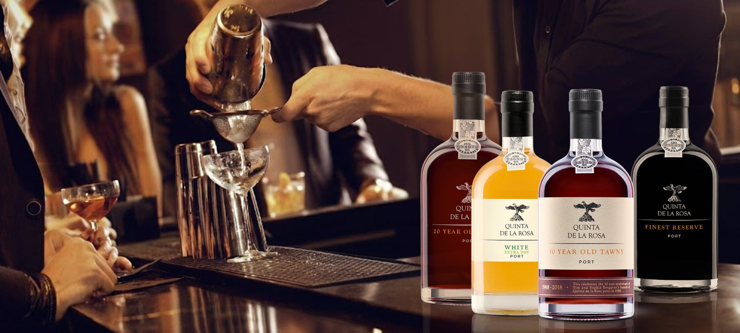 Headerbild für Vinho Bar: Eine Reihe Quinta de la Rosa Portweinflaschen – von hellem White Port bis zum dunklen Ruby Reserve – präsentiert auf einem dunklen Tresen. Im Hintergrund herrscht ein warmes, atmosphärisches Bar-Ambiente mit Gästen und einem Barkeeper.
