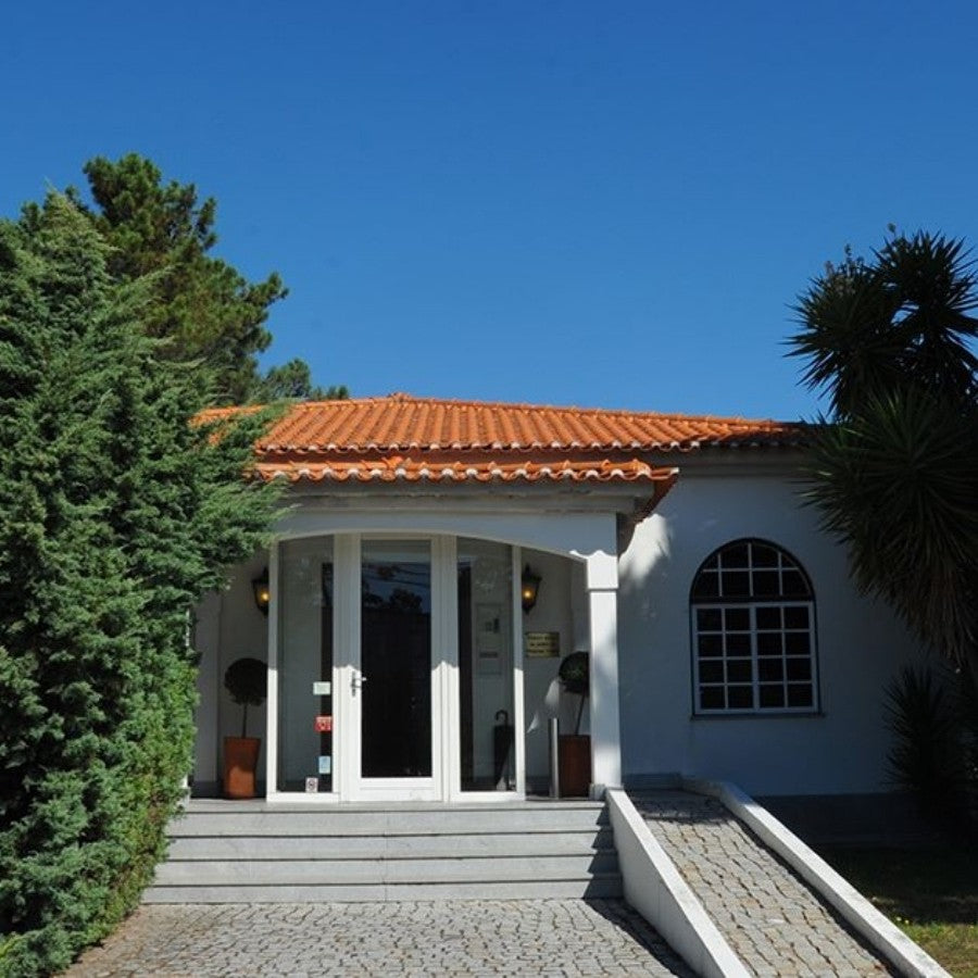 Die Außenansicht des weißen Hauptgebäudes von Quinta de Cabriz in Portugal, umgeben von mediterranen Bäumen und Sträuchern unter blauem Himmel.