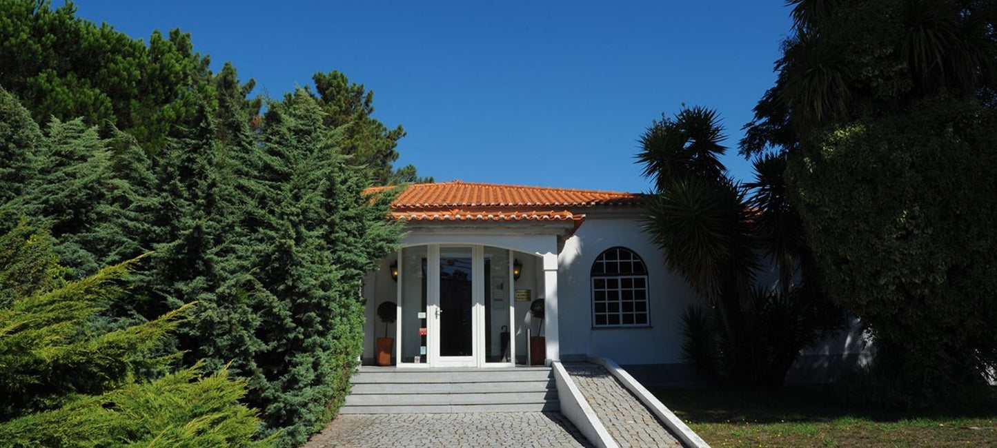 Die Außenansicht des weißen Hauptgebäudes von Quinta de Cabriz in Portugal, umgeben von mediterranen Bäumen und Sträuchern unter blauem Himmel.