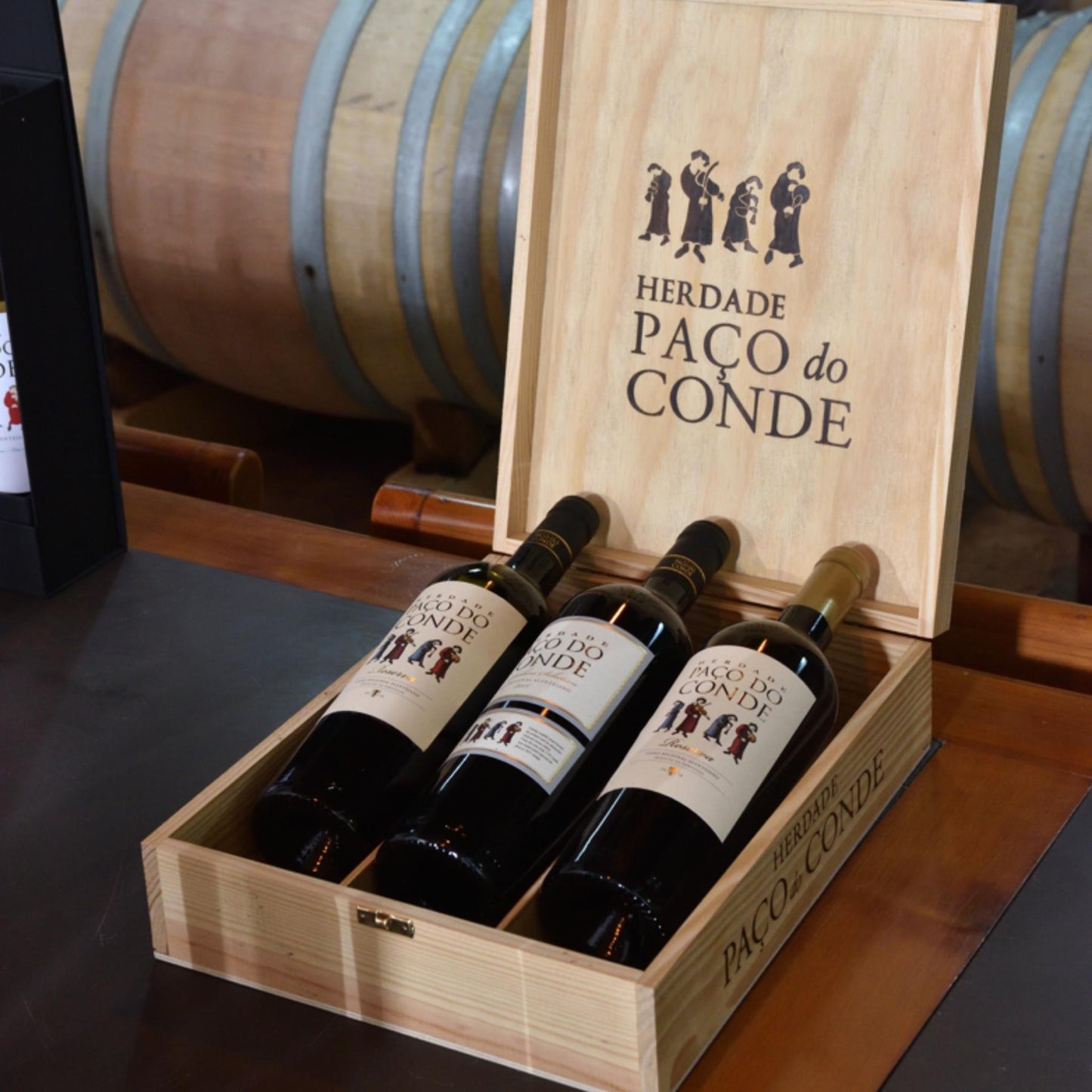 HERDADE PAÇO DO CONDE RESERVA: Rotwein vom Weingut Herdade Paco do Conde aus dem dem Alentejo/Portugal von der Vinho Bar. Drei Weinflaschen in einer Holzkiste mit Logo des Weingutes vor Weinfässern.