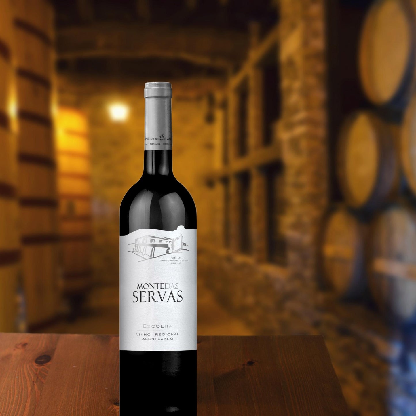 MONTE DAS SERVAS ESCOLHA: Rotwein vom Weingut Herdade das Servas aus der Region Estremoz – Alentejo/Portugal. Flasche in einem Weinkeller auf dem Tisch stehend. Im Hintergrund sind Weinfässer zu sehen.