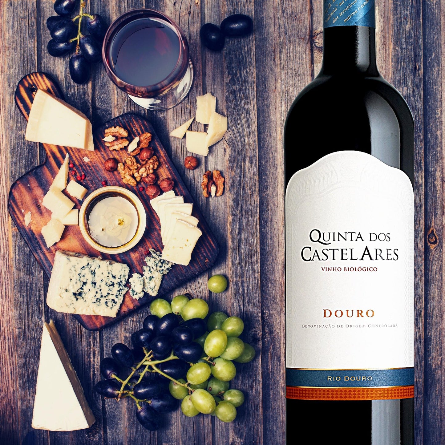 
                  
                    Quinta dos Castelares Colheita Rotwein, Biowein aus dem Douro / Portugal. Bild mit Weinflasche vor einem Holztisch mit Weinglas, Käse auf einem Holzbrett und grünen und roten Weintrauben.
                  
                
