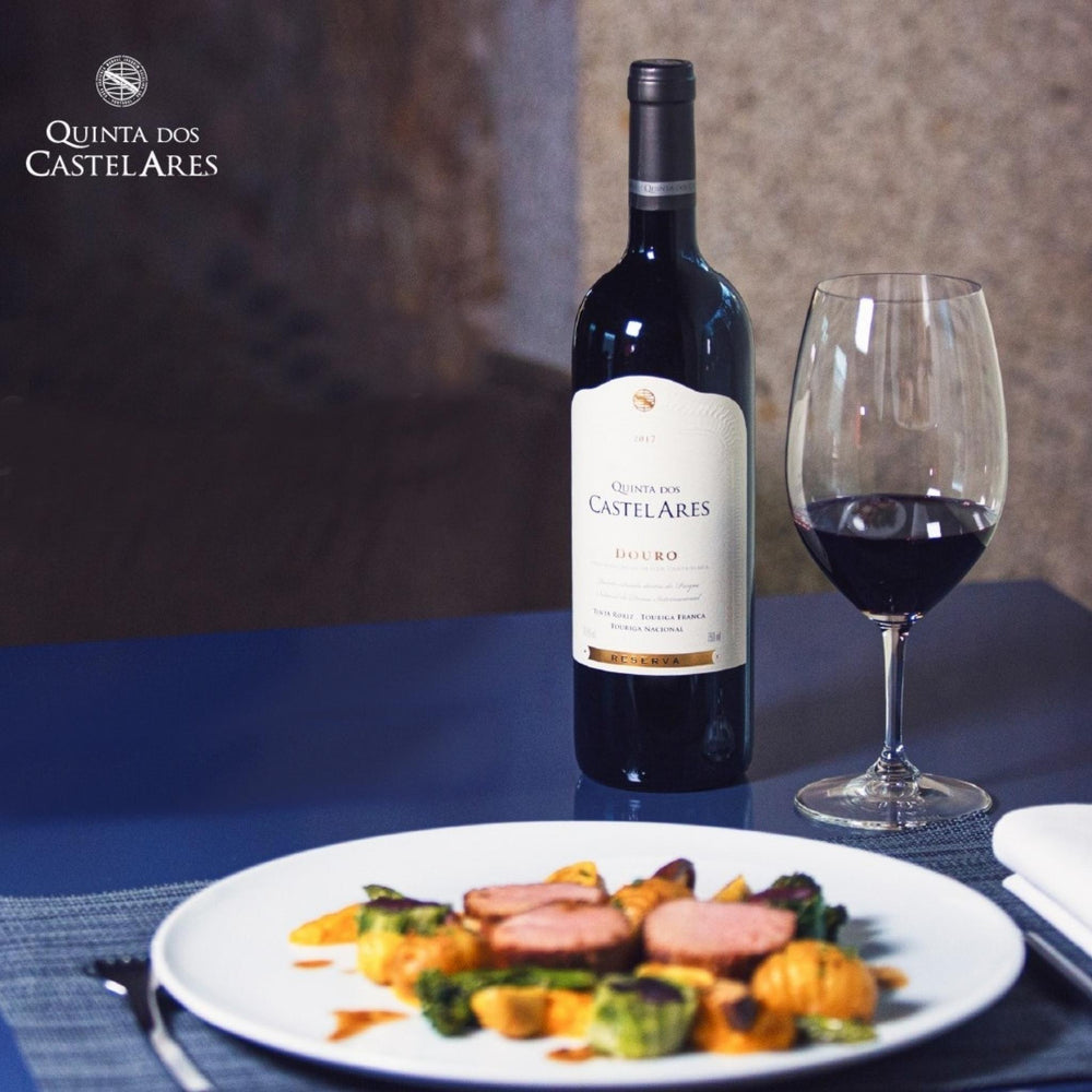 Quinta dos Castelares Reserva, Rotwein aus dem Douro / Portugal. Bild mit der Weinflasche auf einem blauen Tisch mit einem gefüllten Rotweinglas und einem Teller mit Fleisch und Gemüse.