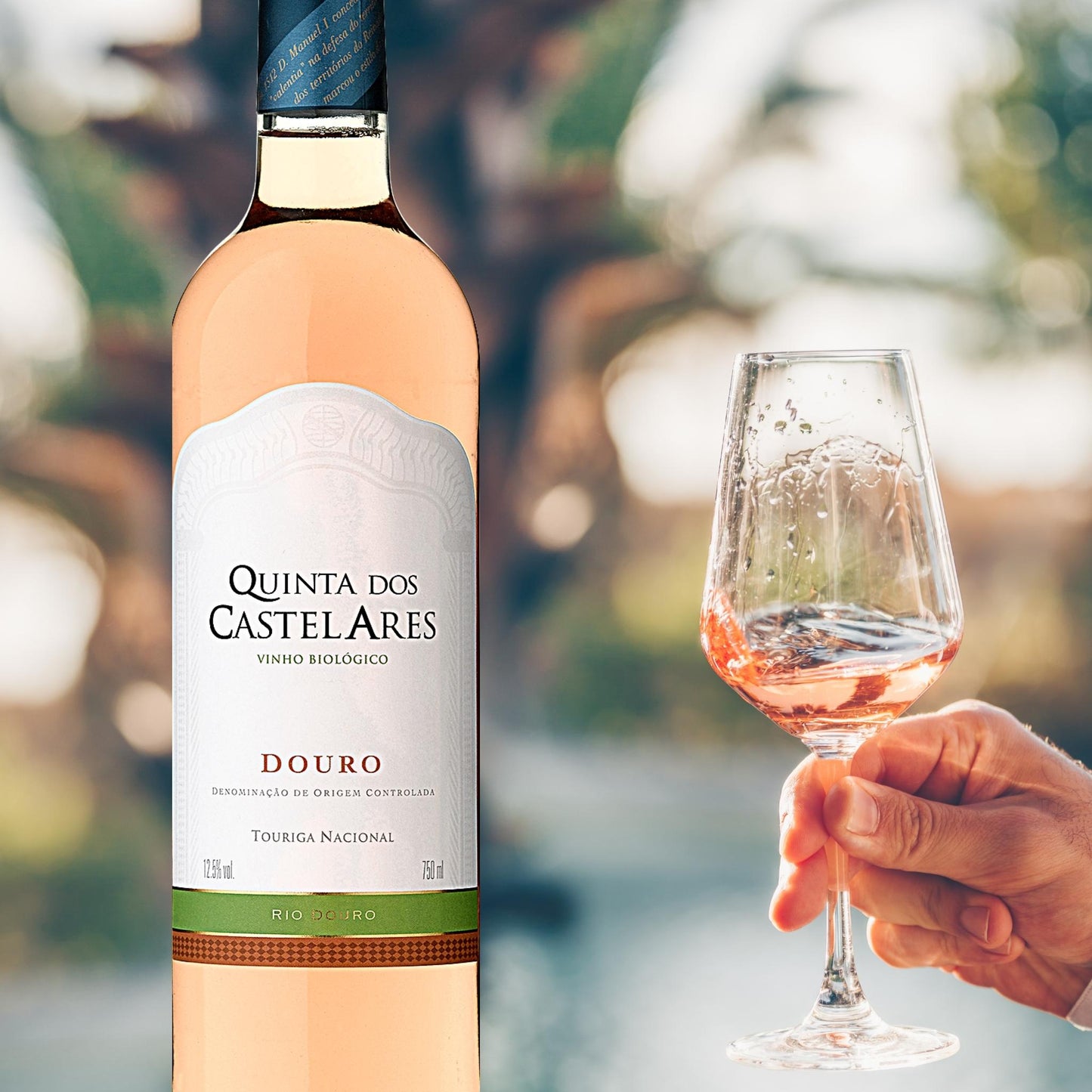 
                  
                    Quinta dos Castelares,  Touriga Nacional 2022 Roséwein / Biowein aus dem Douro / Portugal.  Bild mit Weinflasche und einem Weinglas mit Roséwein. Das Glas wird von einer Männerhand gehalten und geschwenkt.
                  
                